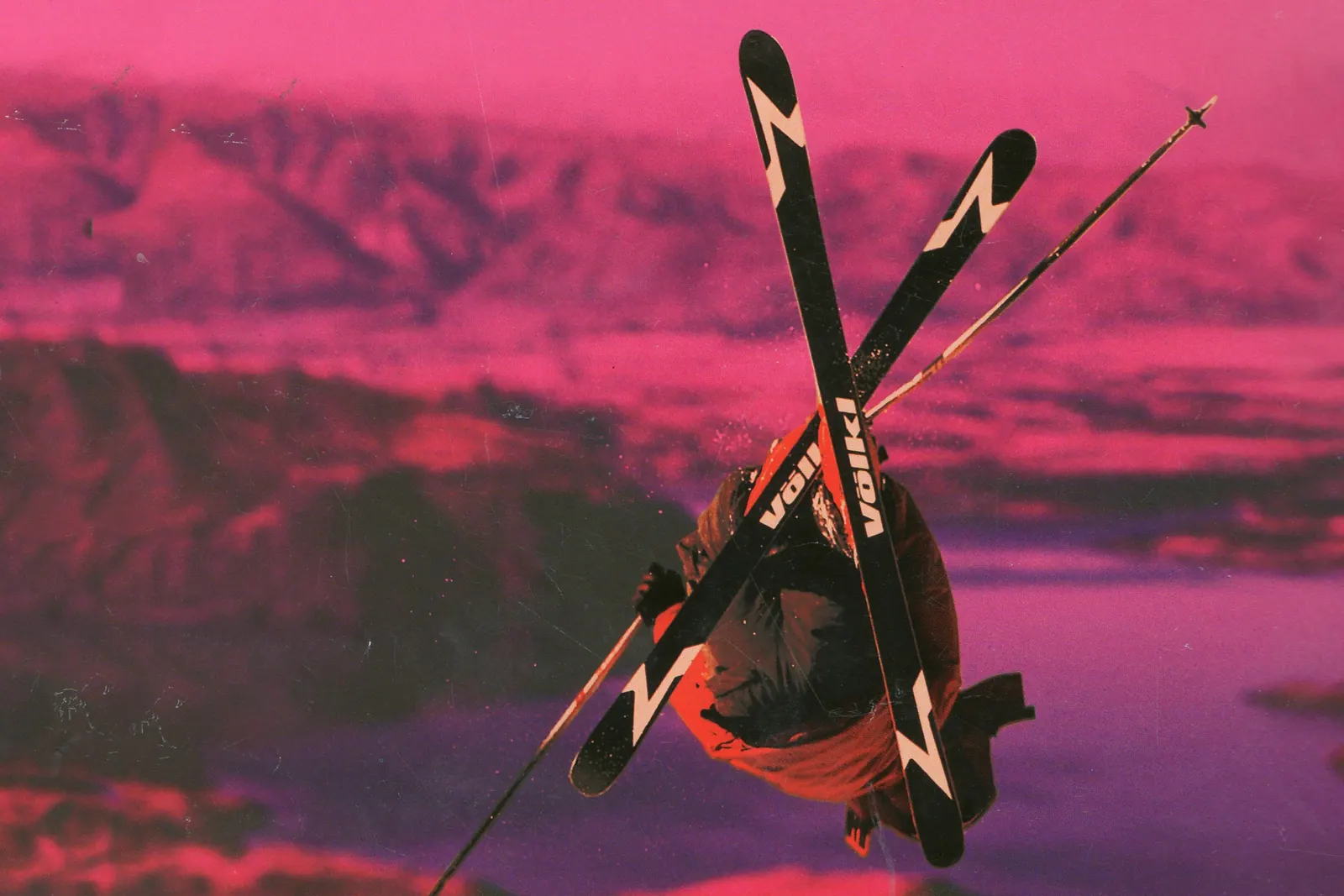 Skifahrer im Sprung mit gekreuzten Völkl-V-Ski vor einer Bergkulisse