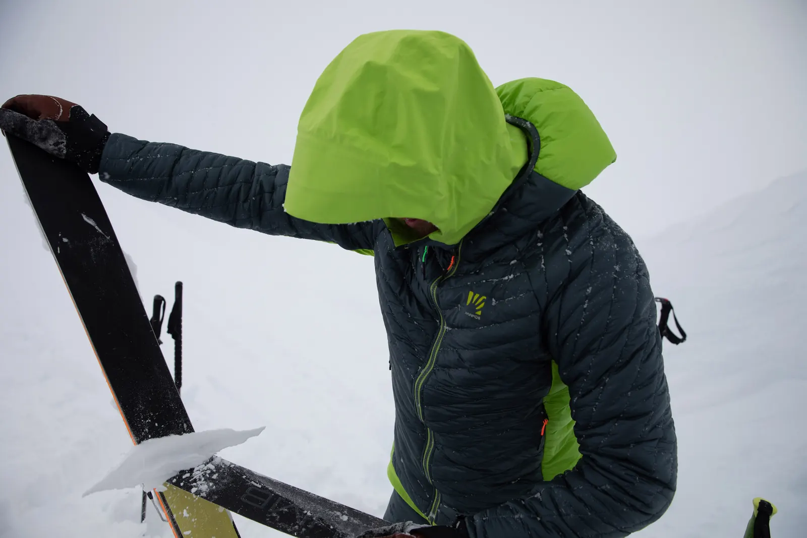 Person mit dunkler Isolationsjacke und heller Kapuze beim Hantieren mit einem Ski im Schnee