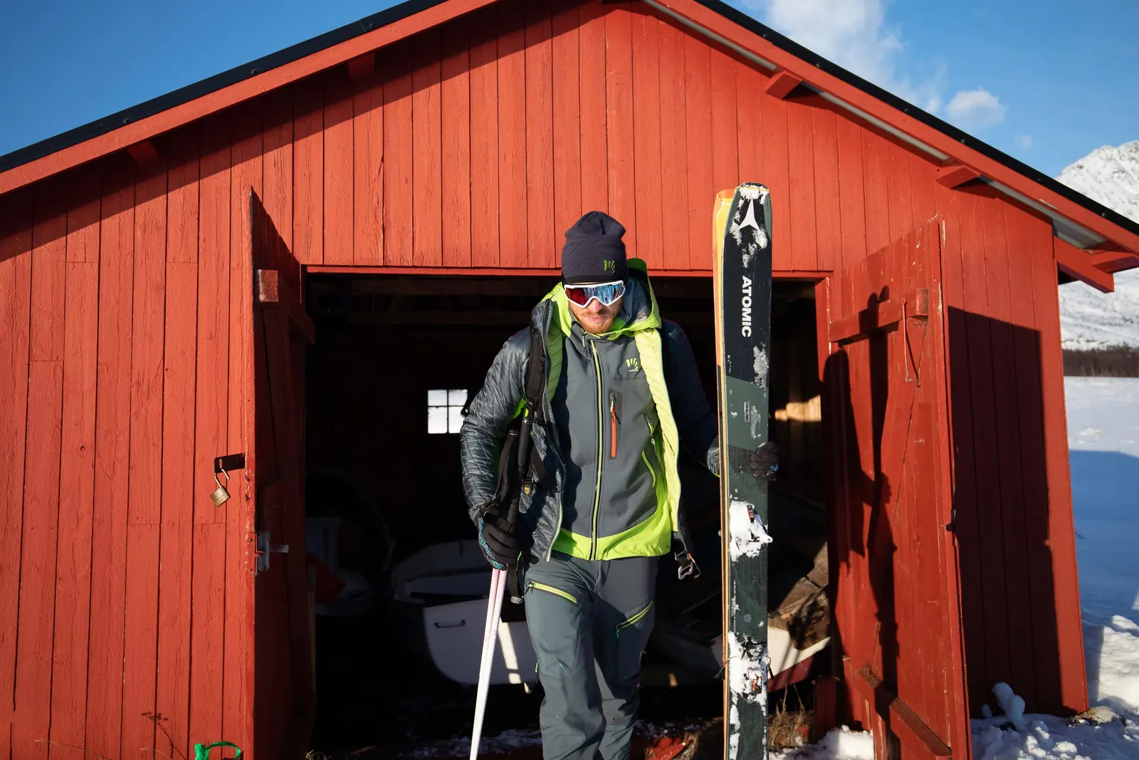 Person mit dunklem Skitourenoutfit und Ski vor einer roten Berghütte