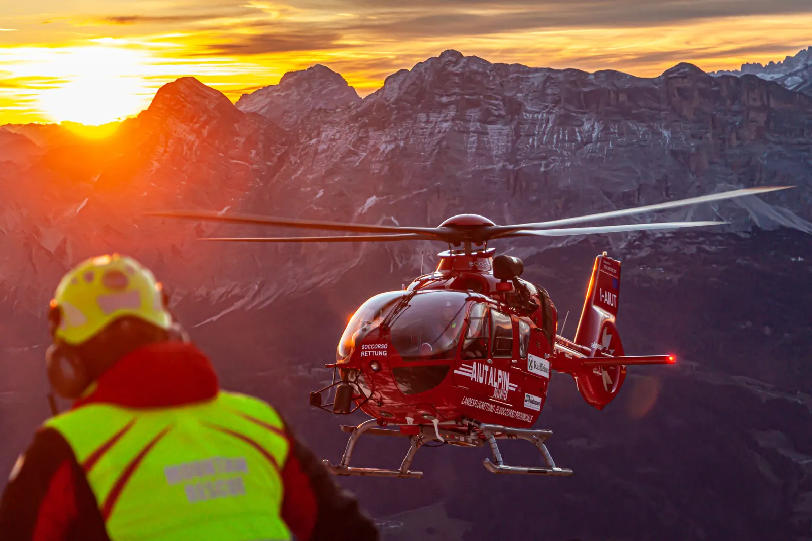 Rettungskraft in auffälliger Einsatzkleidung vor einem Hubschrauber in den Bergen