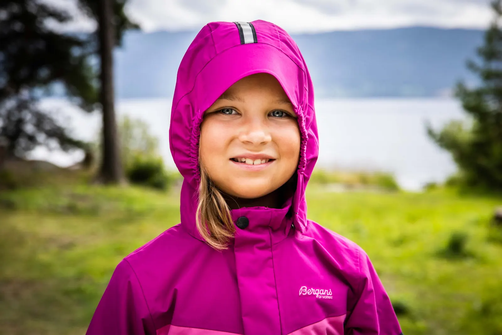 Kind mit geschlossener Kapuze einer violetten Bergans-Jacke vor einem See