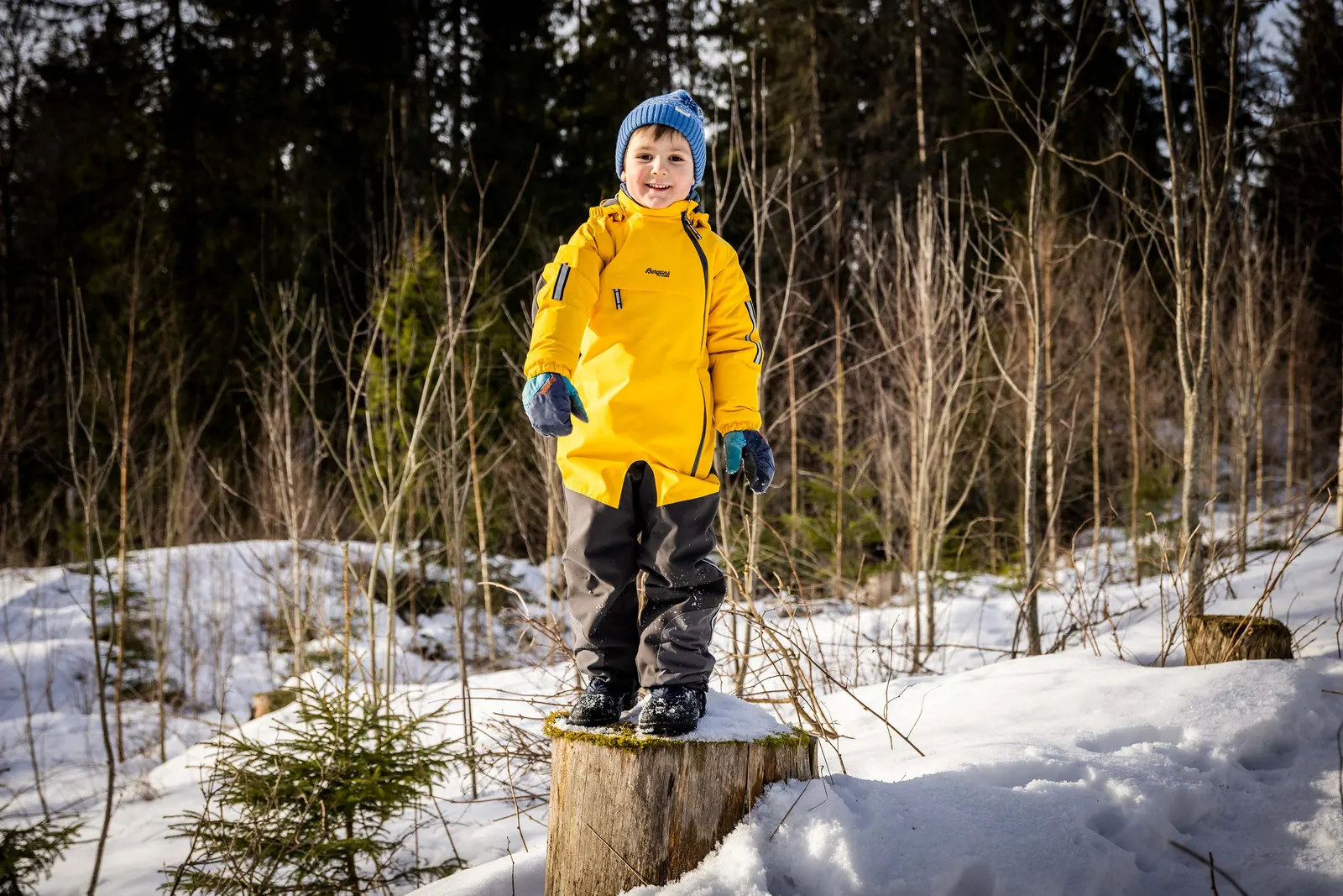 Kind in gelbem Bergans-Overall auf einem Baumstumpf im verschneiten Wald
