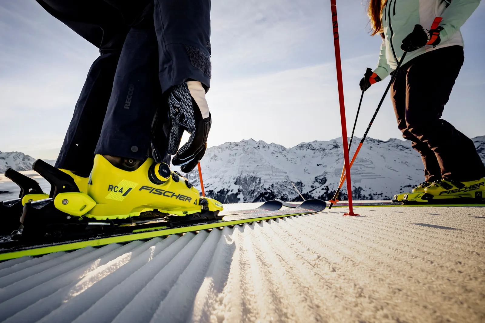 Eine Person stellt den BOA-Verschluss eines gelben Fischer-Skischuhs auf einer präparierten Piste ein
