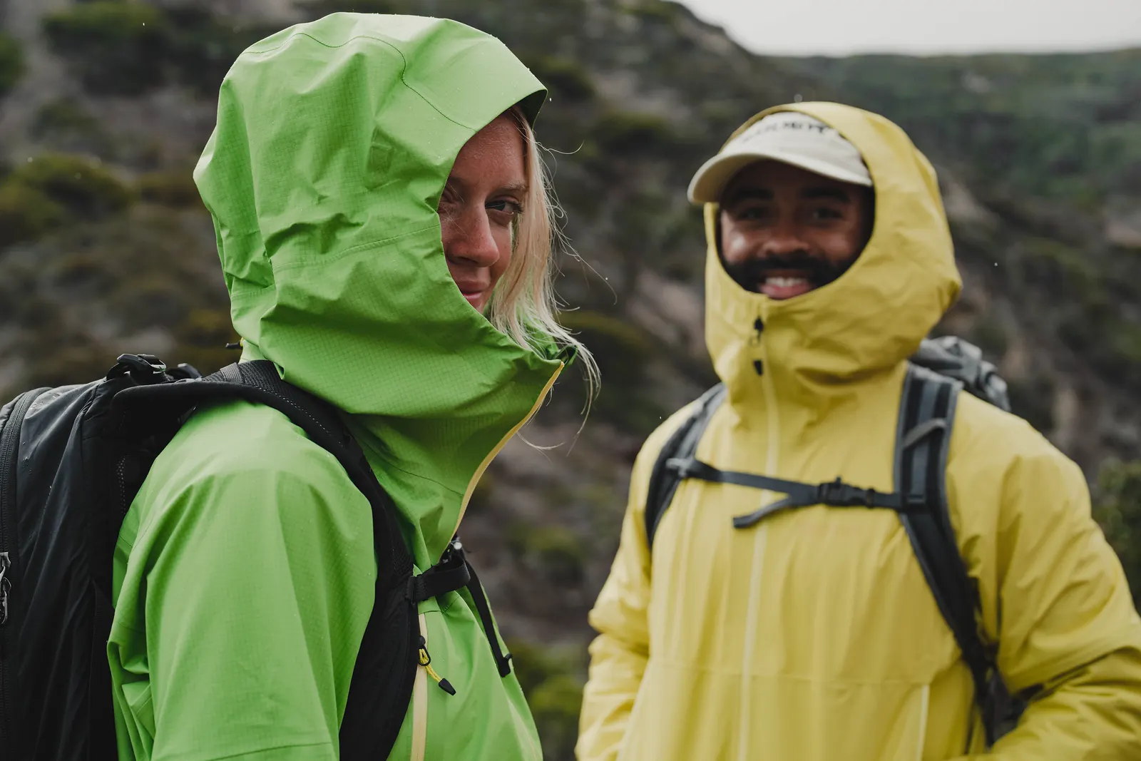 Zwei Personen mit Rucksäcken und geschlossenen Kapuzen an grüner und gelber Hardshelljacke