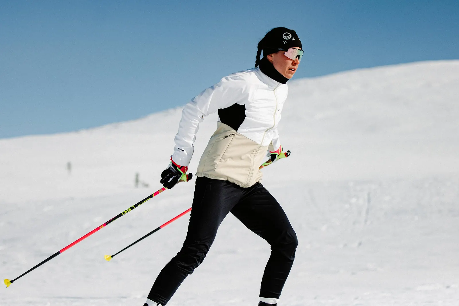 Langläuferin in heller Halti Jacke und schwarzer Hose mit Stöcken auf einer Schneefläche