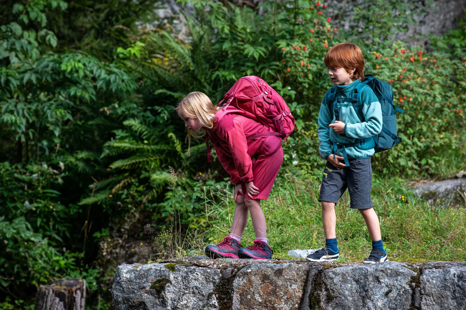 Zwei Kinder mit Rucksäcken stehen in Outdoorbekleidung auf einer Steinmauer