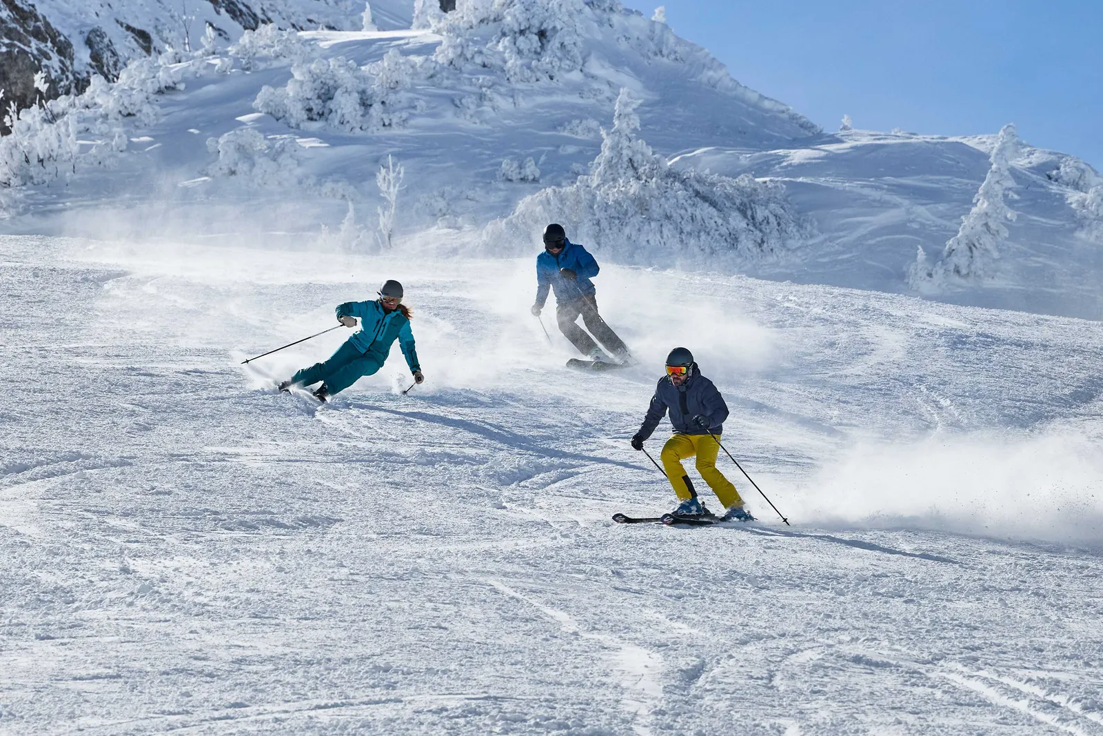 Drei Skifahrende mit türkisen, blauen und gelben Kleidungsstücken auf einer verschneiten Piste