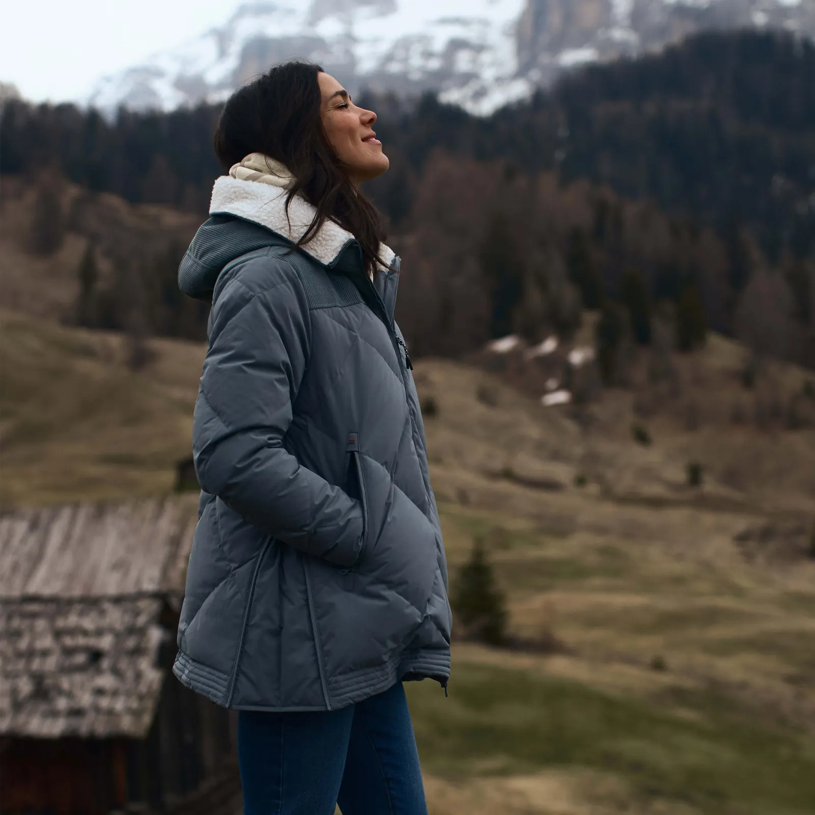 Frau in einer längeren graublauen Dolomite-Jacke vor einer Berglandschaft