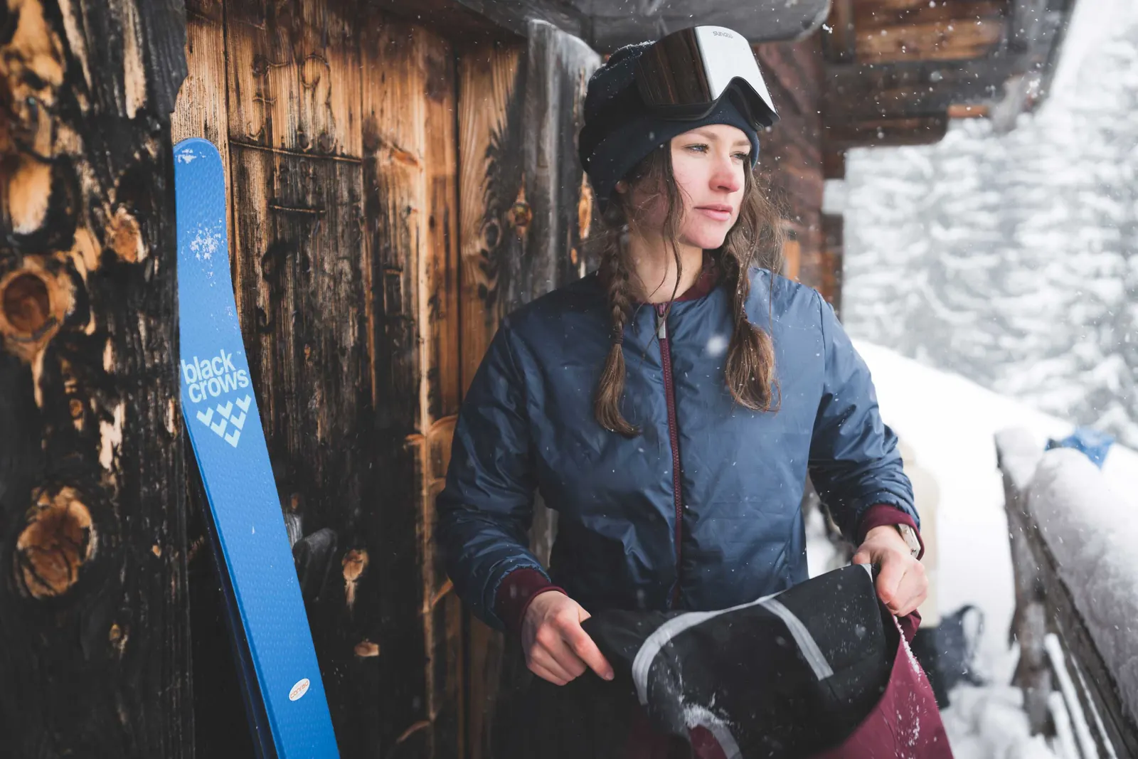 Frau in blauer PYUA Bomberjacke mit elastischen Bündchen neben einer Holzhütte und angelehnten Ski