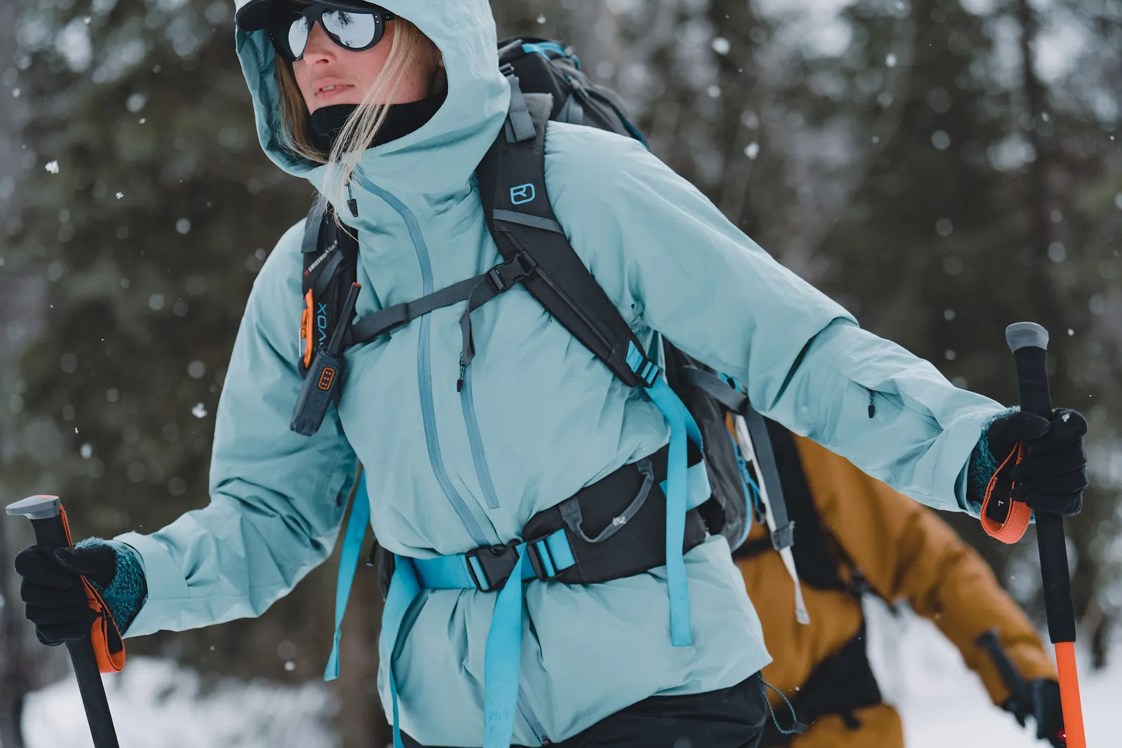 Wintersportlerin mit hellblauer Kapuzenjacke, Rucksack und Stöcken im Schnee