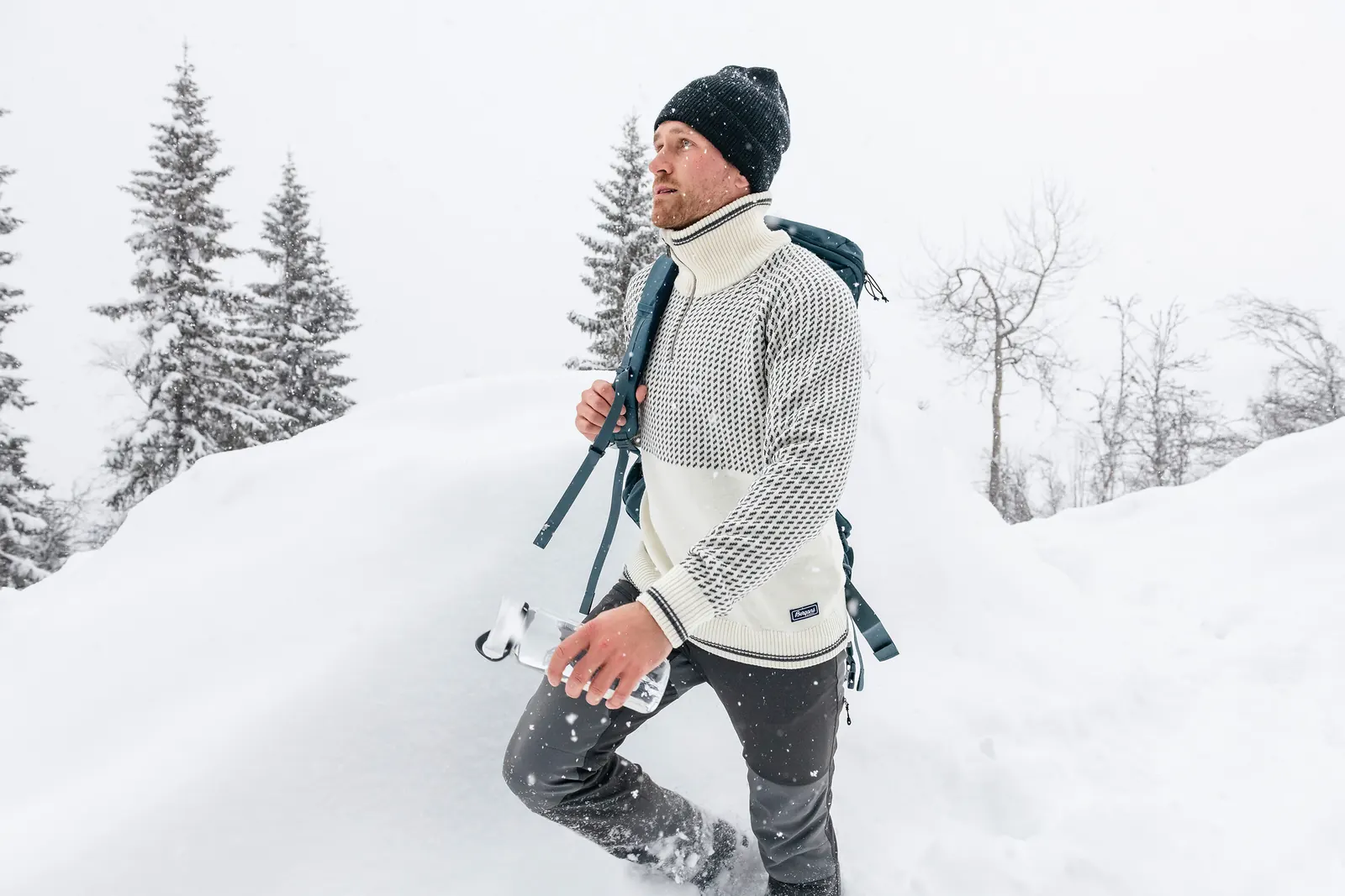 Mann im hellen Strickpullover von Bergans mit geschulterten Skiern im Schnee