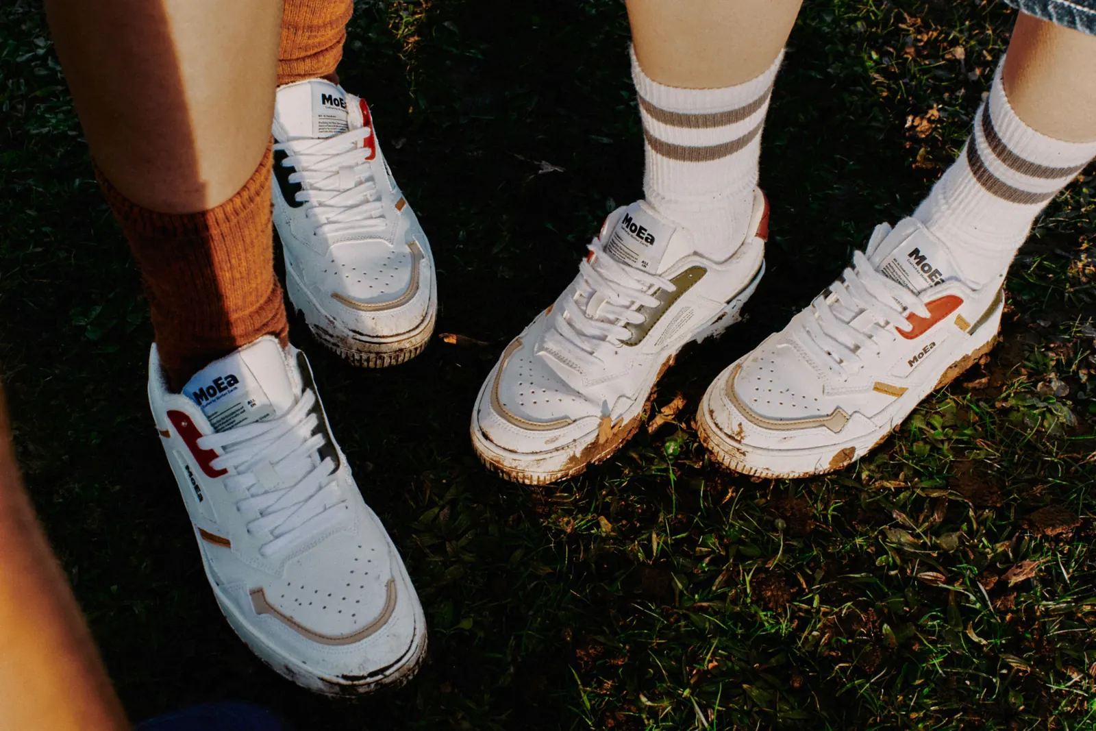 Weiße MoEa-Sneaker mit farbigen Details an den Füßen zweier Personen auf einer Wiese