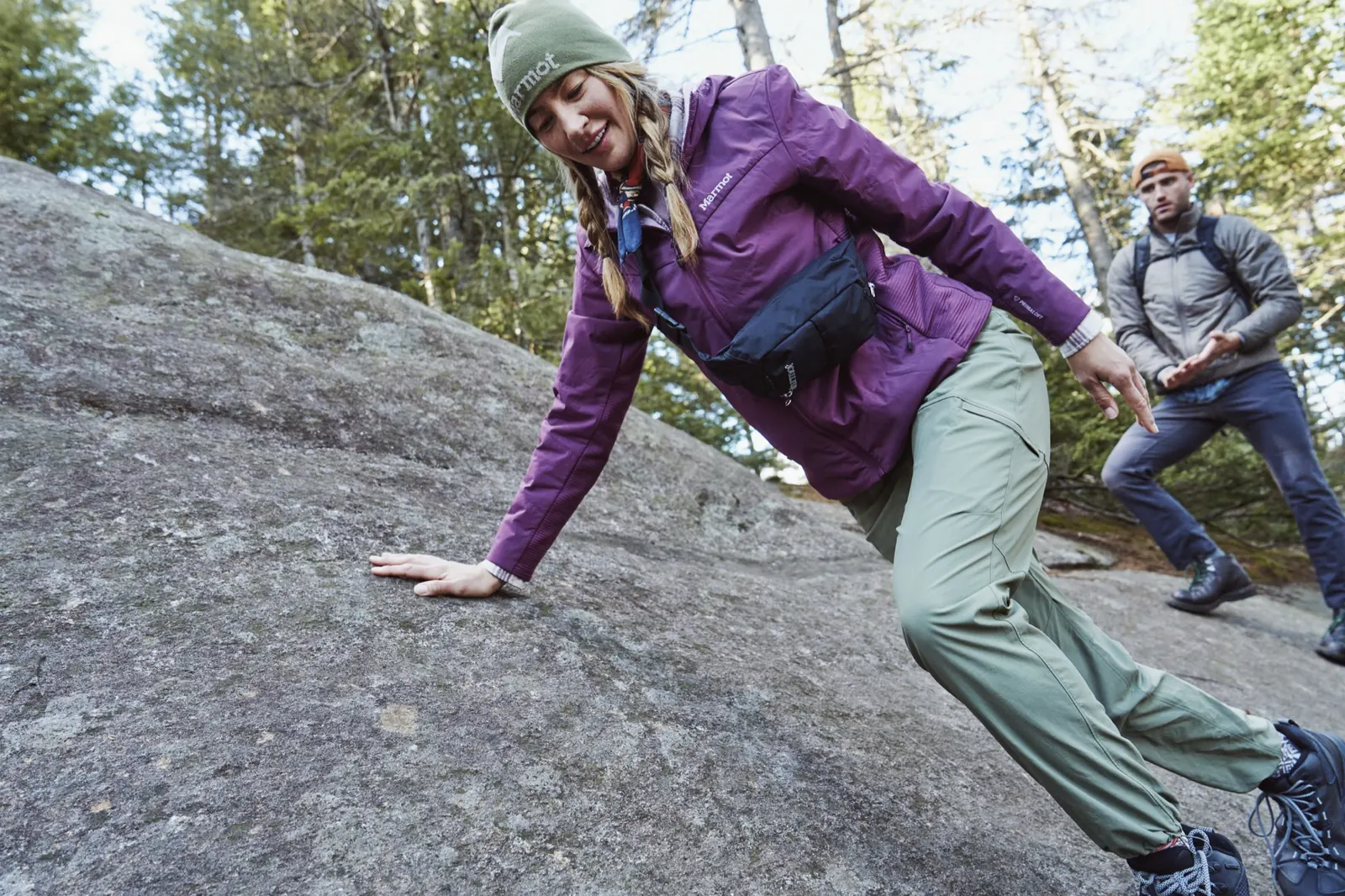 Frau in violetter Marmot Revlo Hybrid Jacket klettert einen Felsen hoch, Mann folgt im Hintergrund