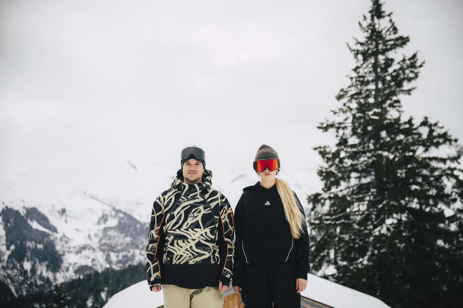 Snowboarder in der O'Neill FWC'Play Asymmetric Snow Jacket und Snowboarderin im O'Neill Hoodie vor Bergkulisse