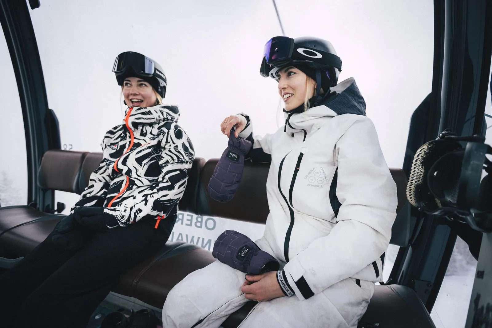 Snowboarderin in der O'Neill FWC'Play Asymmetric Snow Jacket und eine Begleiterin in der Gondel auf dem Weg zum Gipfel