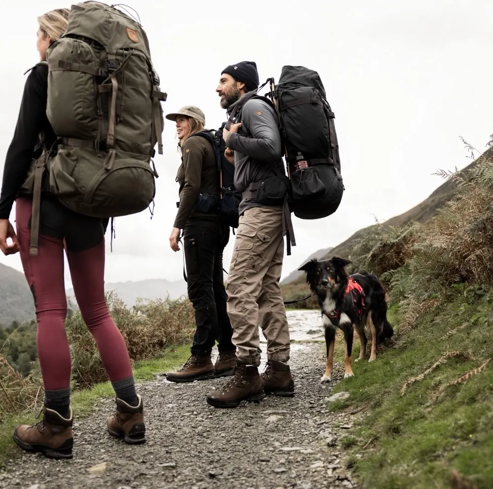 Drei Wandernde mit großen Rucksäcken und ein Hund auf einem nassen Schotterweg im Hochland