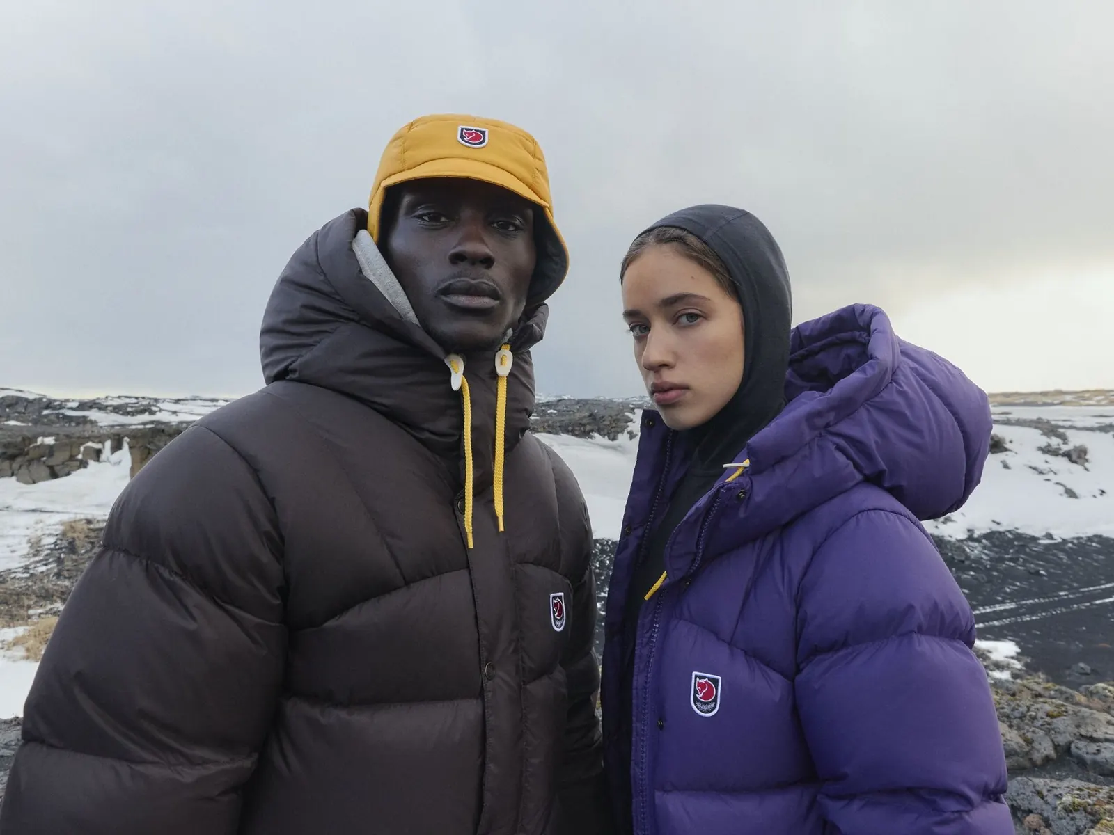 A couple in Fjällräven Expedition Down Lite Jackets, him in dark brown with a yellow beanie, her in violet with the hood up