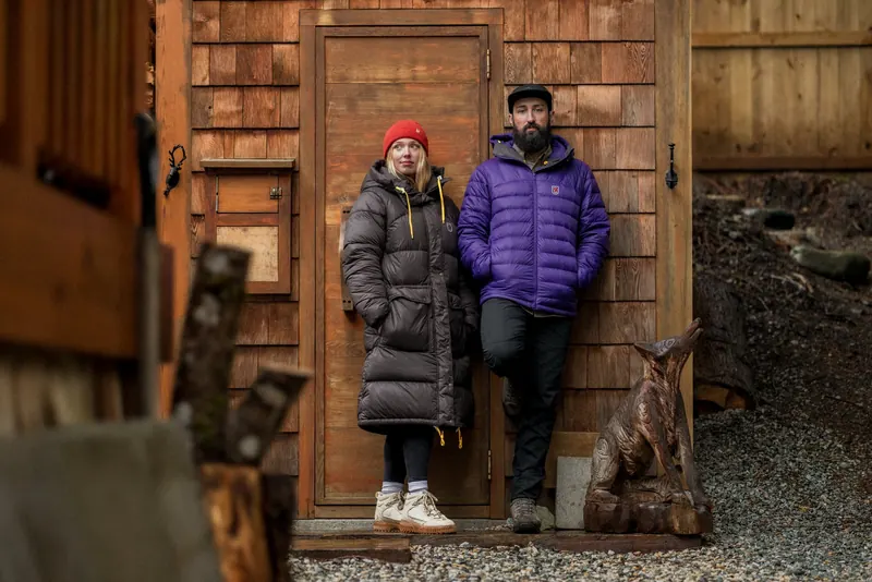 Woman in a long black down jacket and man in a violet Fjällräven down jacket in front of a wooden hut