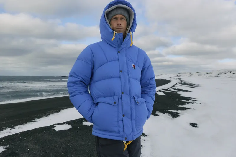 Mann in einer hellblauen Fjällräven Daunenjacke mit Kapuze am schwarzen Strand