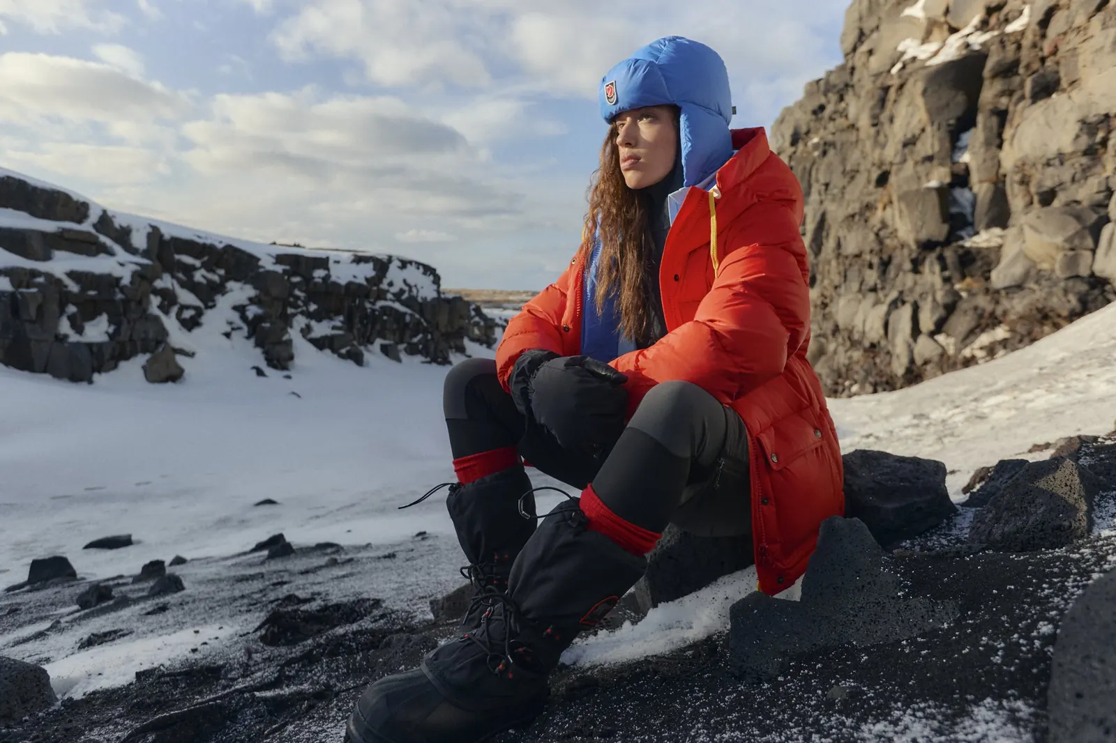 Frau in einer roten Fjällräven Expedition Daunenjacke mit blauer Mütze sitzt auf verschneitem Lavagestein