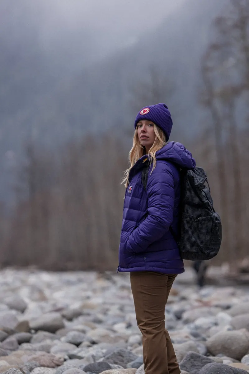 Wanderin in violetter Fjällräven Daunenjacke mit Rucksack in einem Flussbett im Nebel