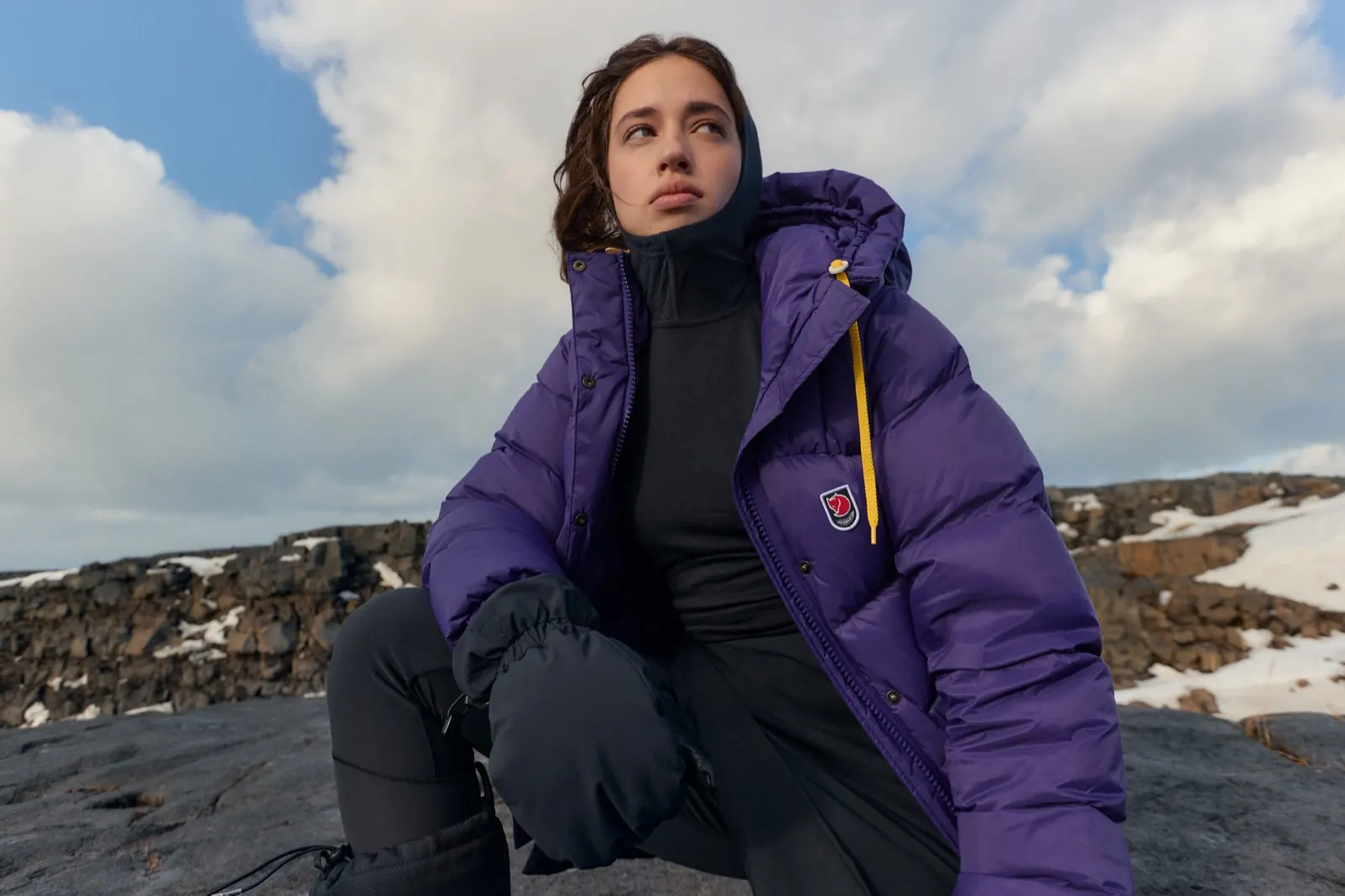 Frau in einer lila Fjällräven Expedition Daunenjacke mit Knopfleiste und gelber Kordel hockt auf einem Lavafelsen