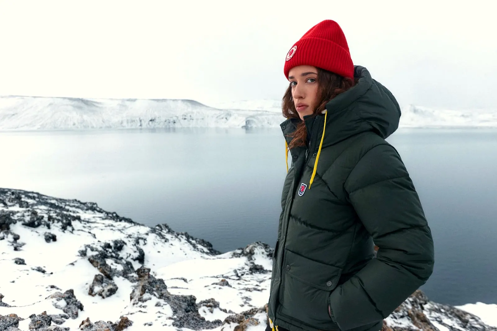 Frau mit roter Mütze in einer dunkelgrünen Fjällräven Expedition Daunenjacke an einem verschneiten Kratersee