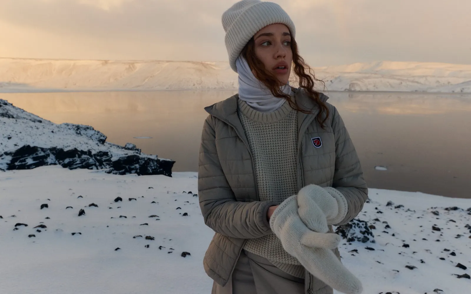 Frau mit heller Mütze und beiger Fjällräven Steppjacke hält Fäustlinge an einem verschneiten See