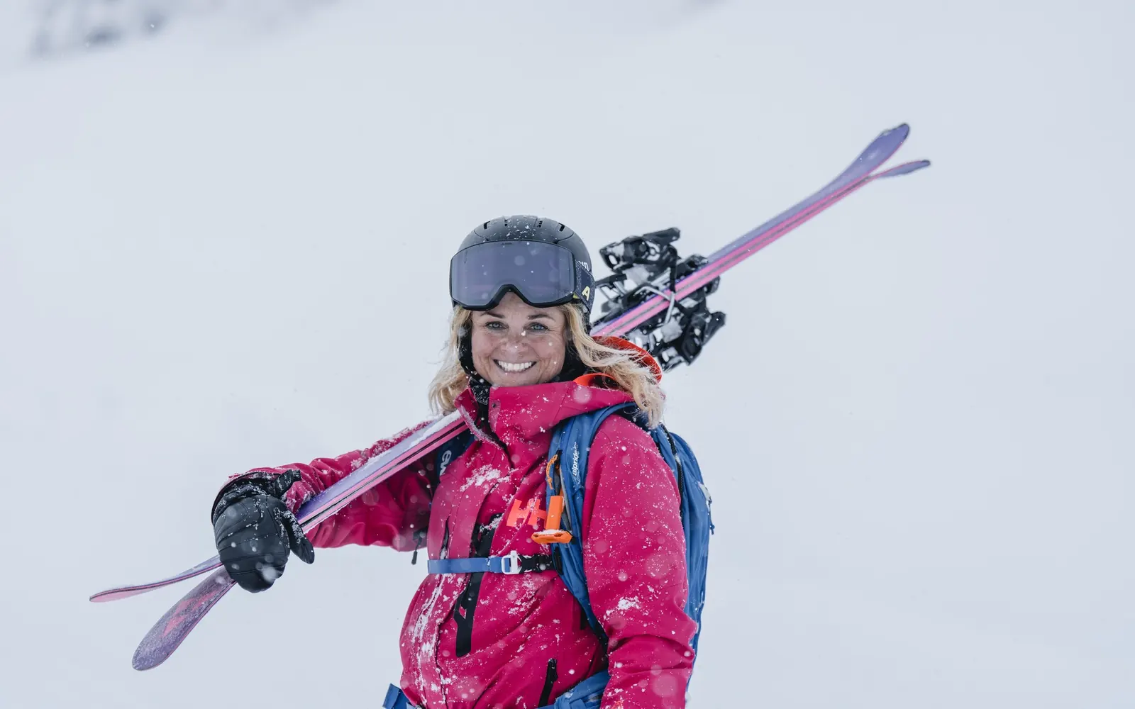 Smiling skier in a pink Helly Hansen jacket carrying her skis on her shoulder