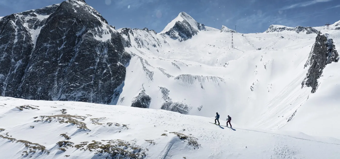 Zwei Tourengeher steigen über einen verschneiten Rücken, dahinter Gletscher und Gipfel des Kitzsteinhorns