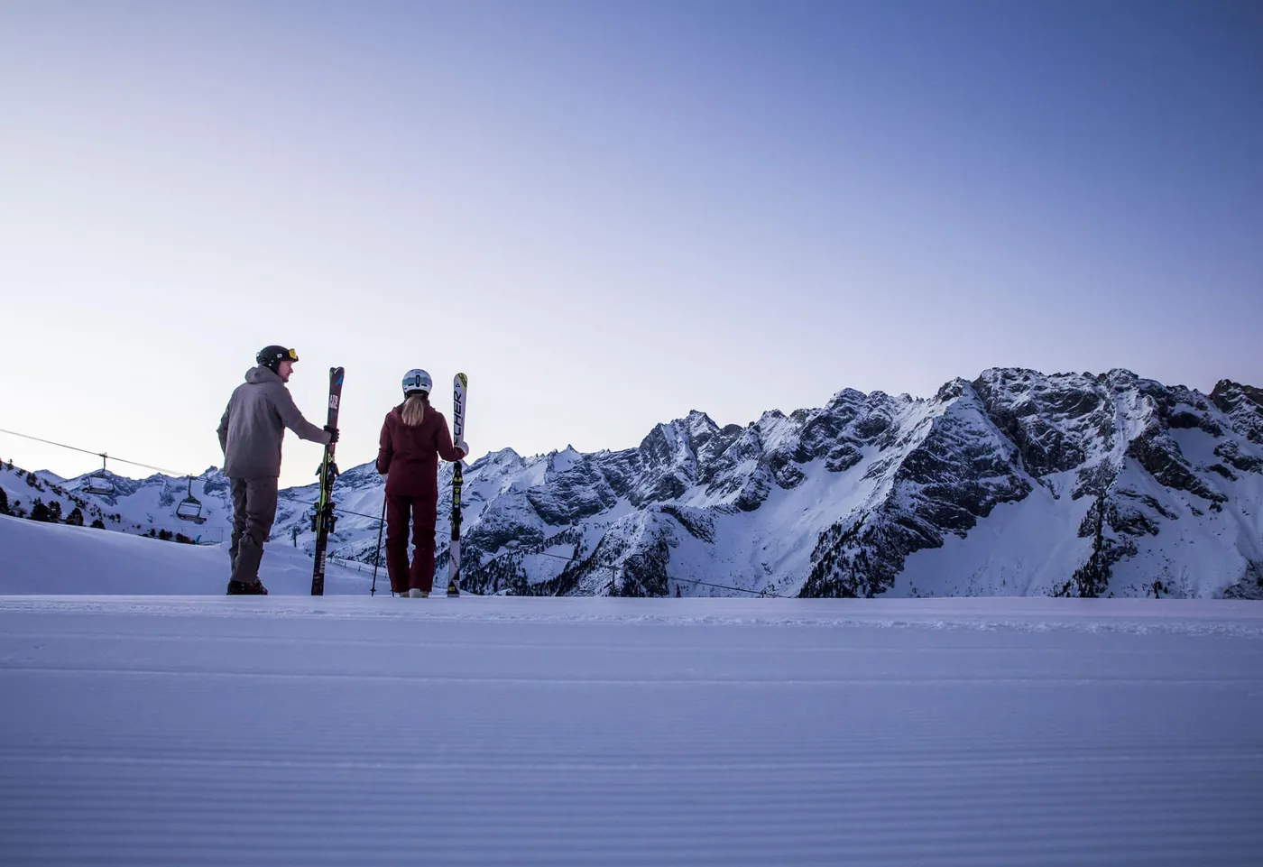 Zwei Skifahrer mit geschulterten Ski auf frisch präparierter Piste, dahinter Felsgipfel