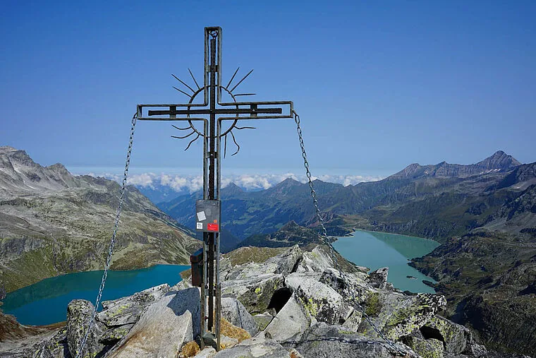 Gipfelkreuz auf einem Felsgipfel, tief unten zwei türkise Stauseen im Stubachtal
