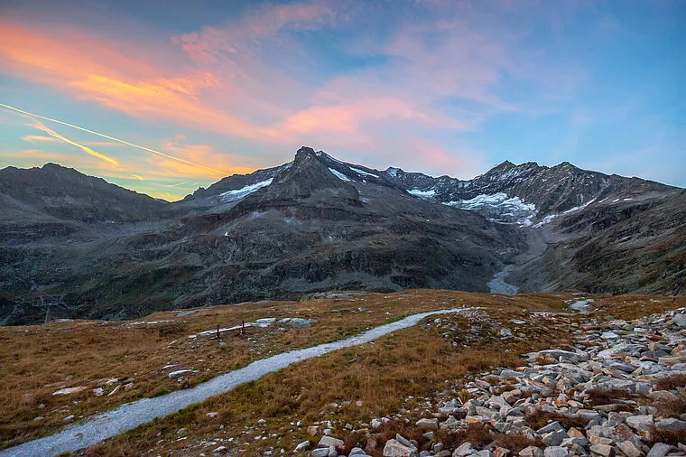 Schmaler Wanderweg über Almwiesen bei der Rudolfshütte, dahinter Gipfel mit Schneefeldern