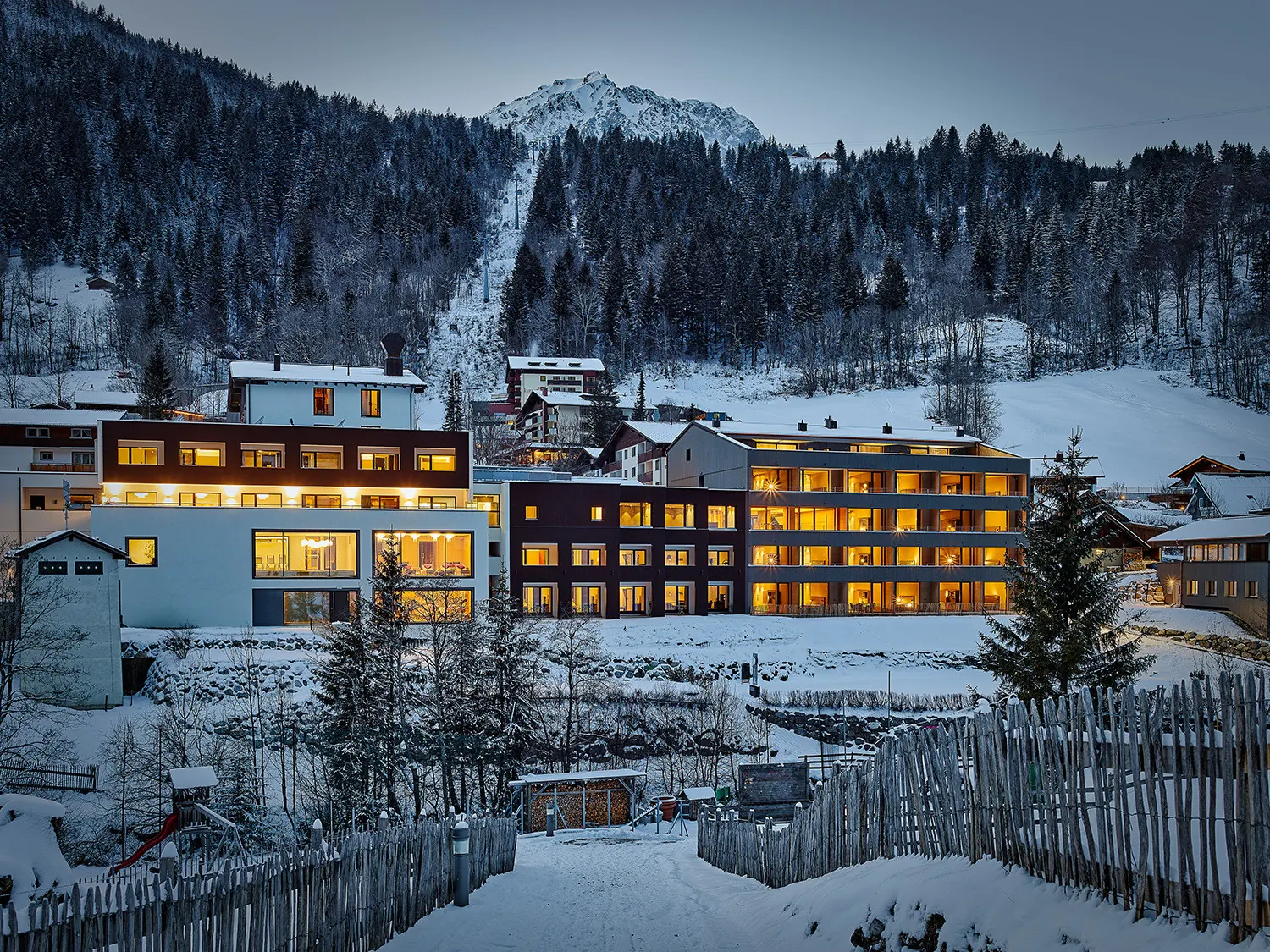 Hotel Sarotla in Brand an einem Winterabend, beleuchtete Fenster, dahinter Wald und die Trasse der Dorfbahn