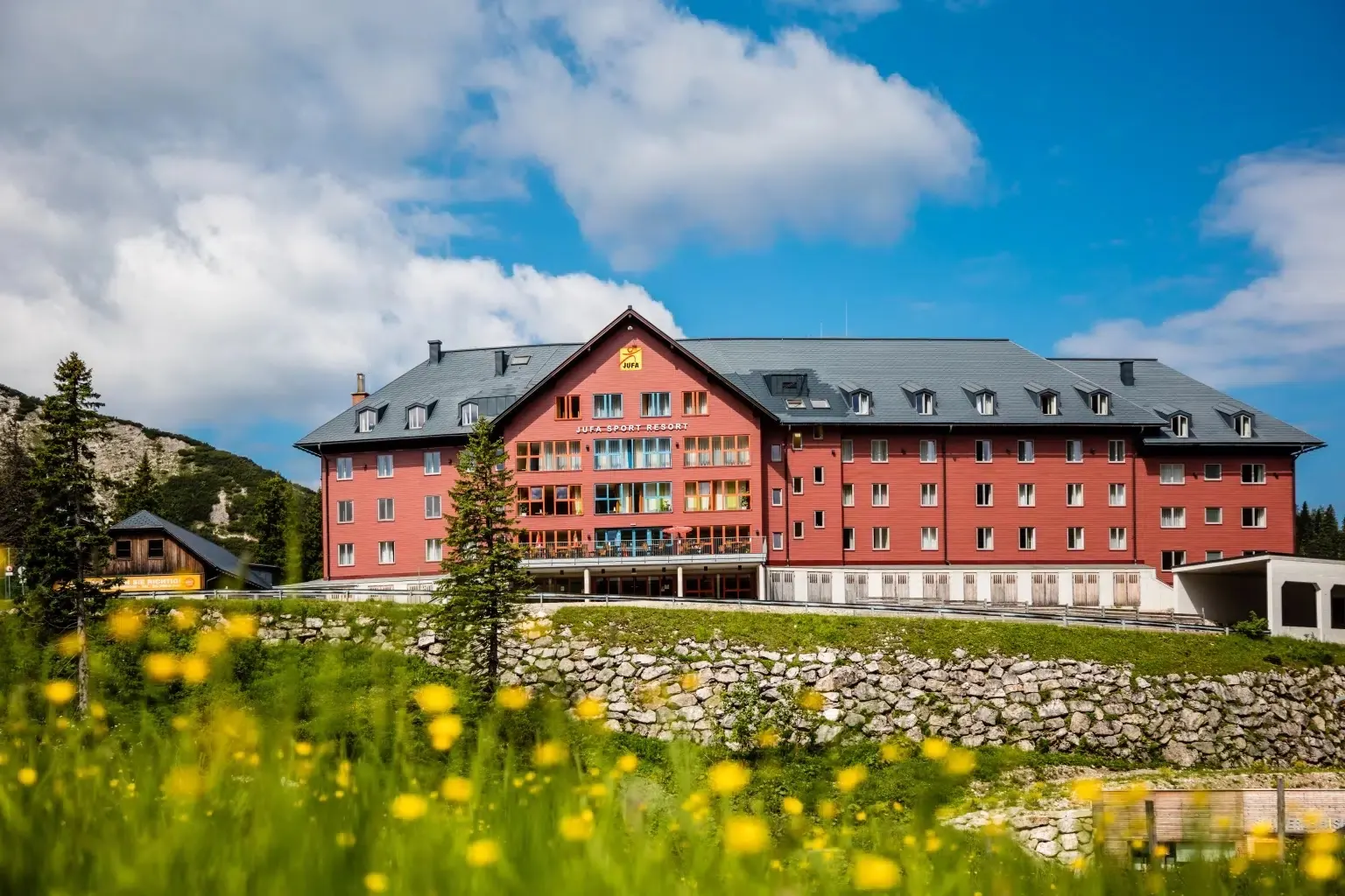 Rote Holzfassade des JUFA Hotel Hochkar mit Schriftzug Sport Resort, davor eine Blumenwiese im Sommer