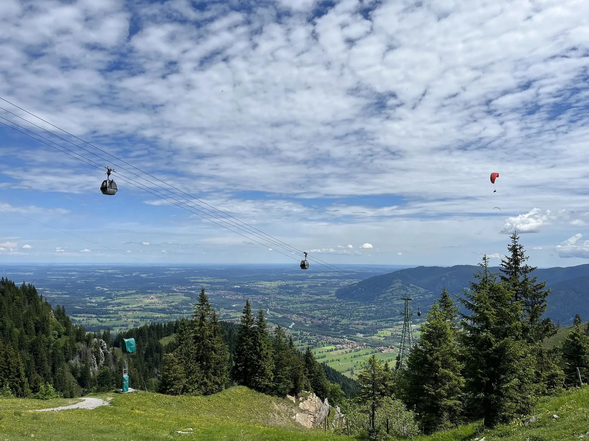 Blick vom Brauneck über grüne Wiesen und Wald ins Isartal mit Gondeln über dem Hang
