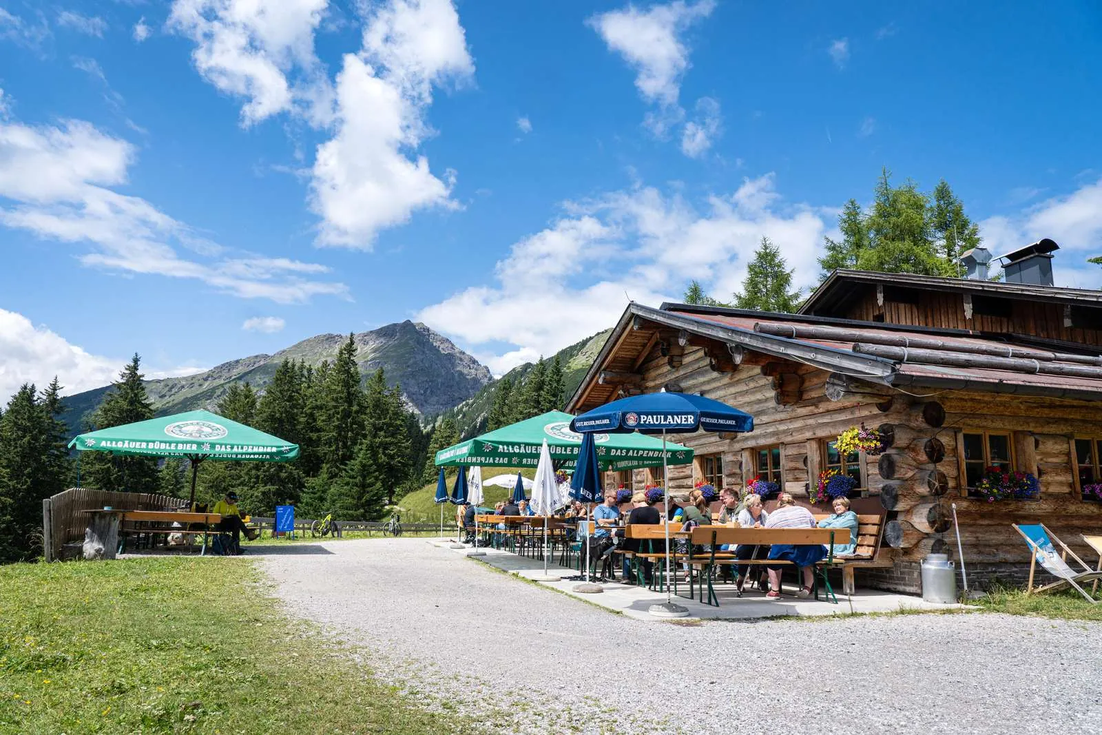 Heiterwanger Hochalm mit besetzter Sonnenterrasse, Wiese und bewaldeten Berghängen im Sommer.
