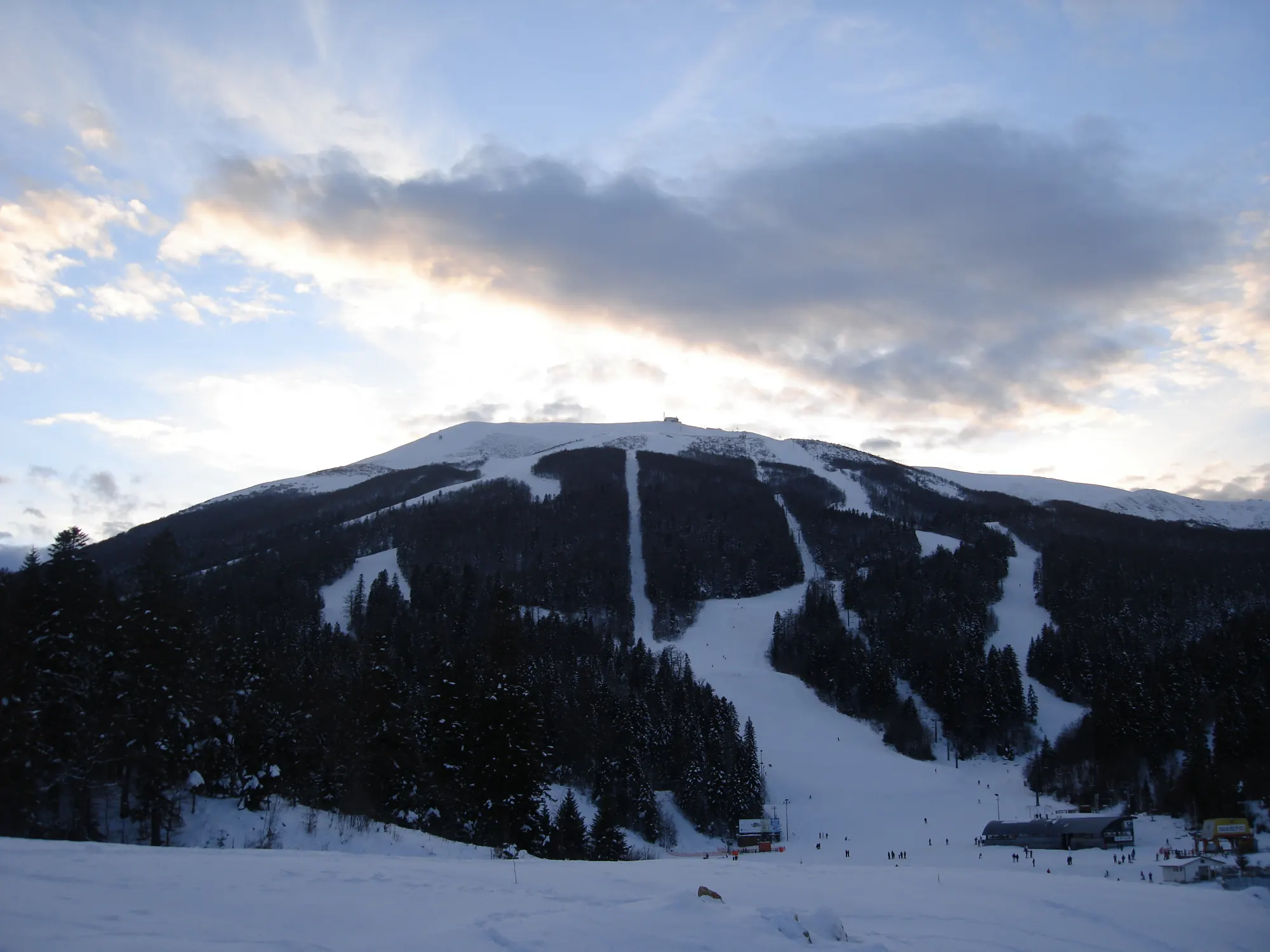 Bewaldeter Berg Bjelašnica mit mehreren Pistenschneisen und Liftanlagen im Abendlicht, unten Skifahrer an der Talstation