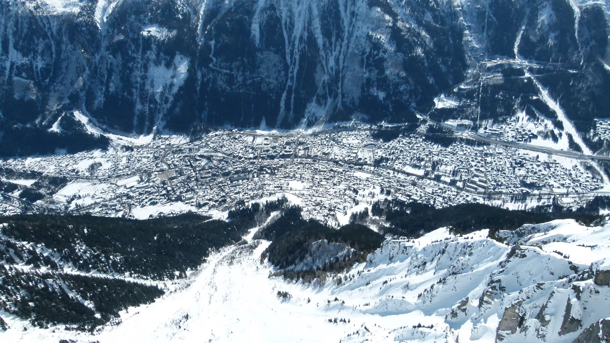Blick vom Brévent hinunter auf das verschneite Chamonix im Tal, gegenüber steile bewaldete Berghänge mit Schneerinnen