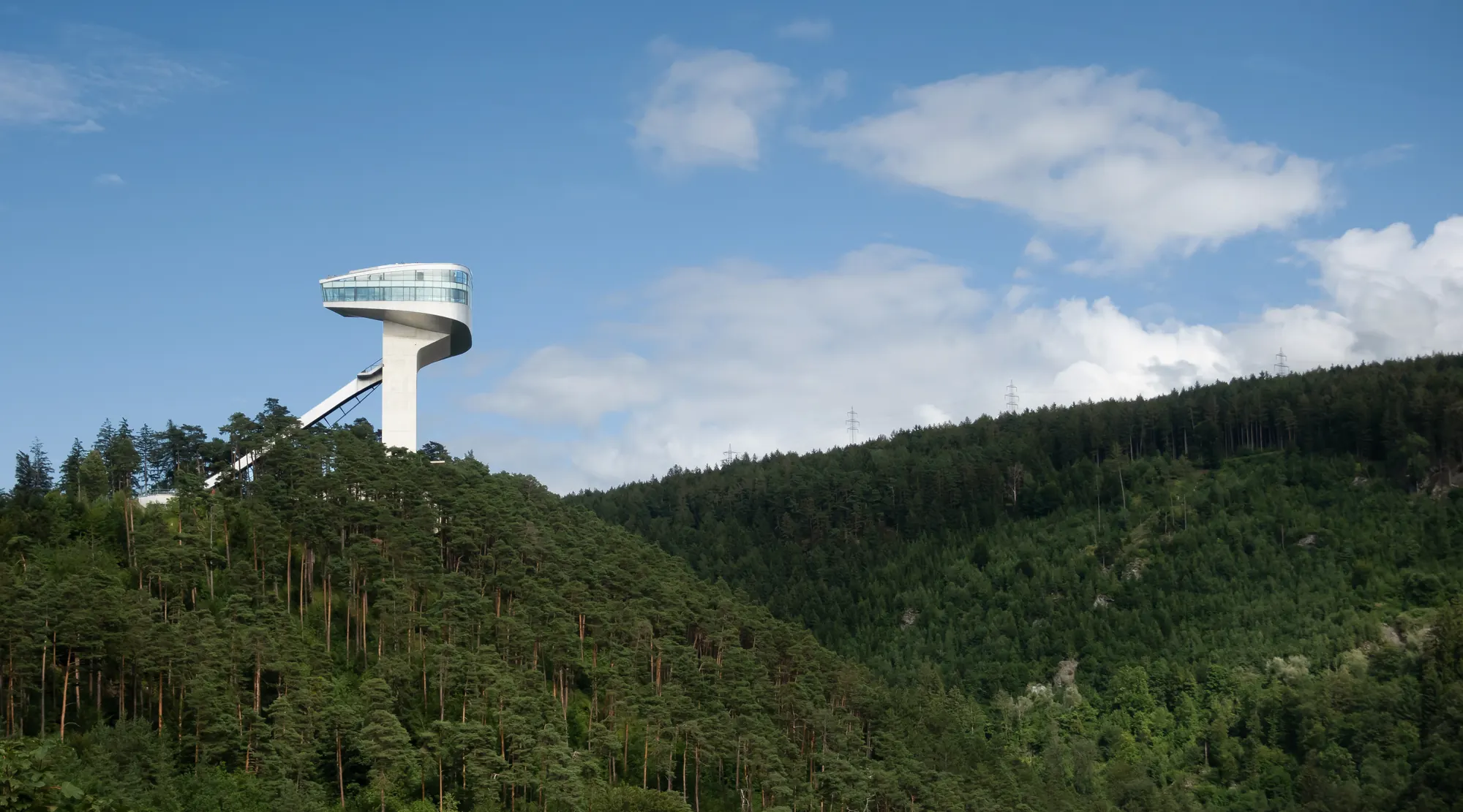 Turm der Bergiselschanze in Innsbruck über bewaldetem Hang, darüber blauer Himmel mit Wolken im Sommer