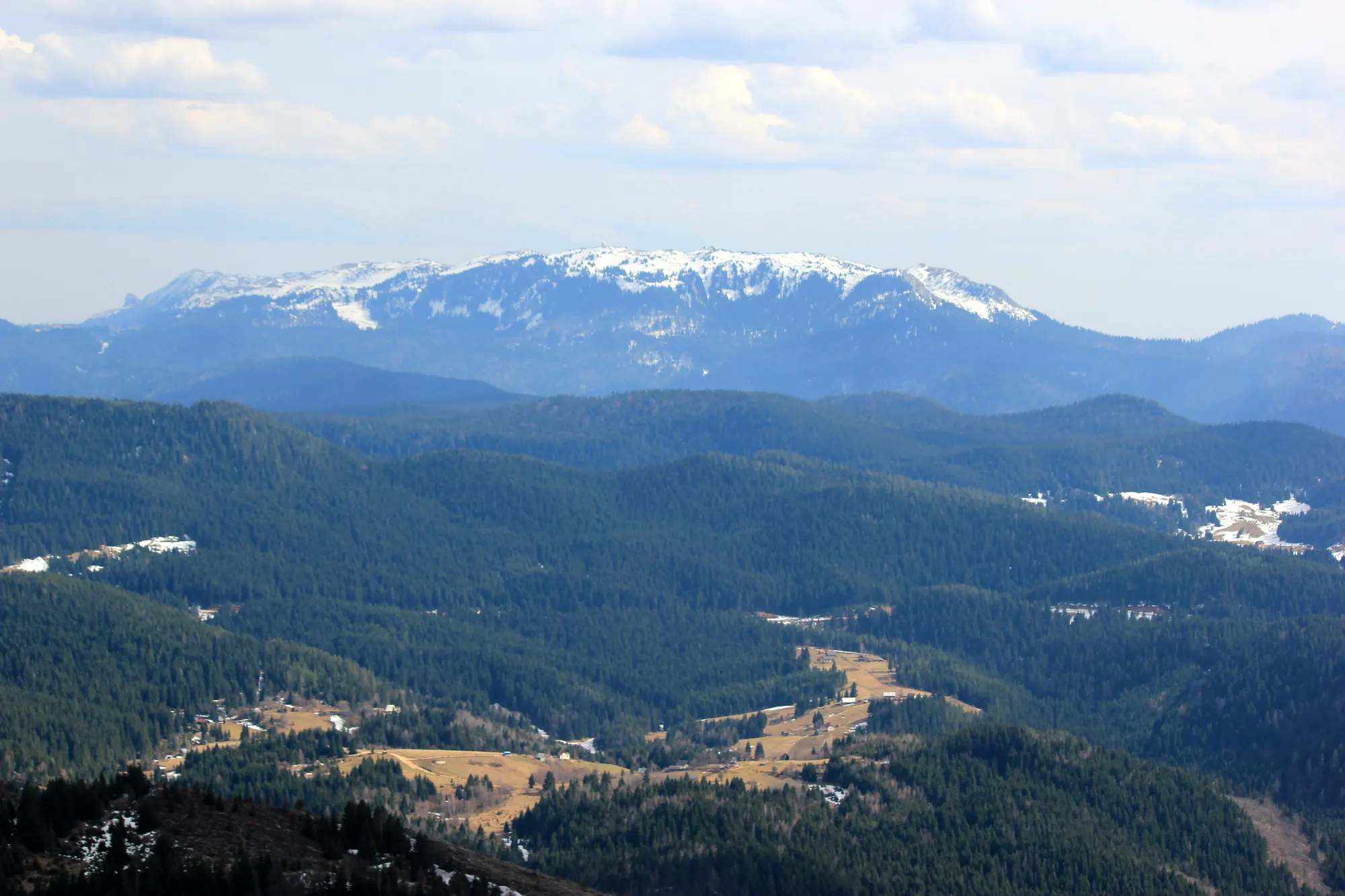 Blick vom Trebević auf die Jahorina mit schneebedecktem Kamm über bewaldeten Hügeln und Lichtungen