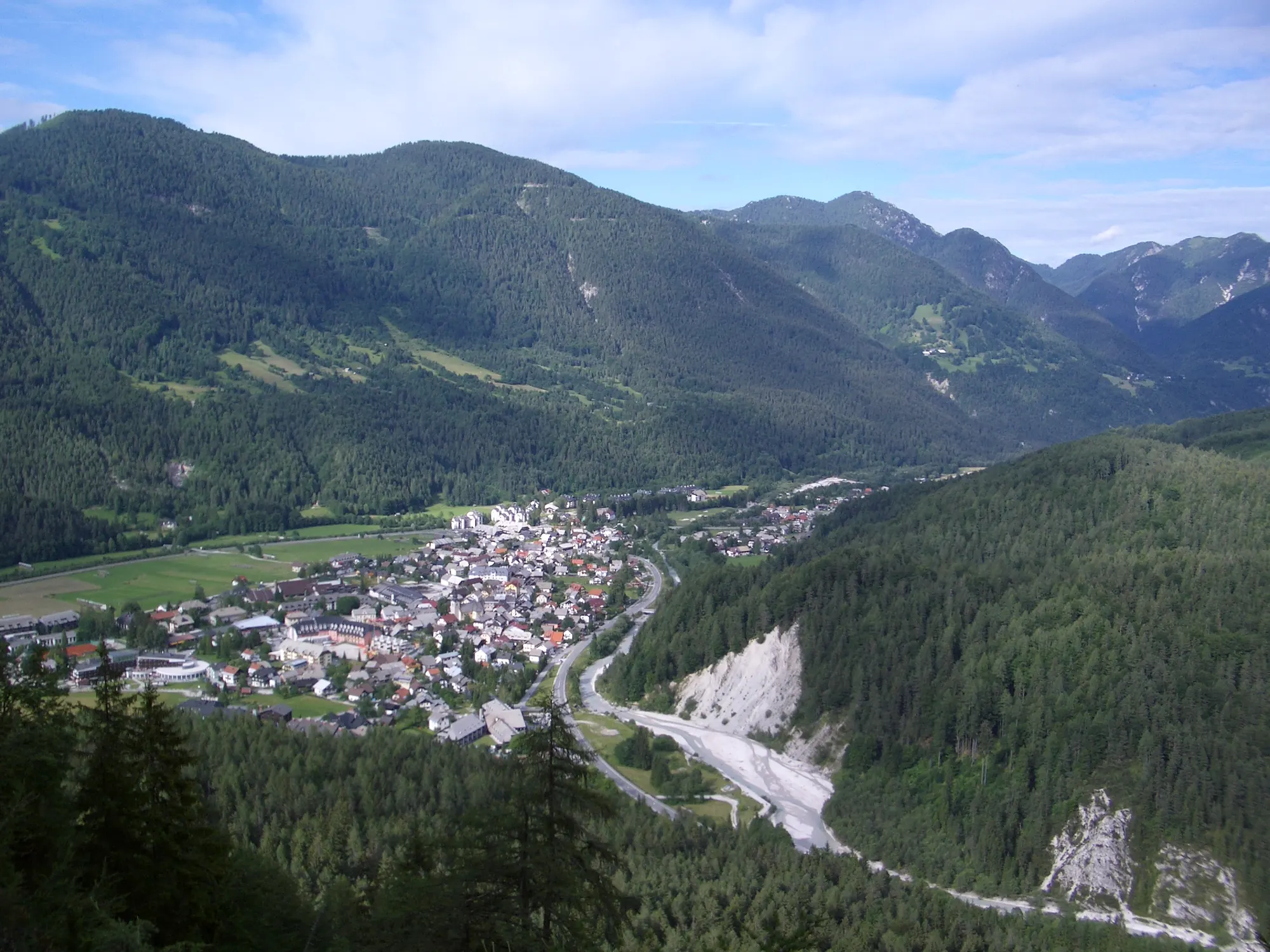 Blick vom Vitranc auf Kranjska Gora im Sommer, mit Ort im Talboden, Flussbett und bewaldeten Bergen ringsum