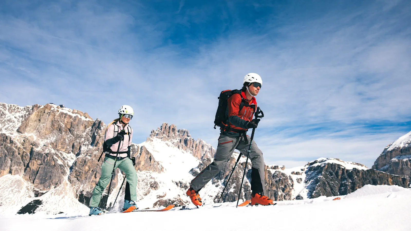 Zwei Skitourengeher mit Helm im Aufstieg vor Dolomitengipfeln, vorne ein Mann in roter Karpos-Jacke