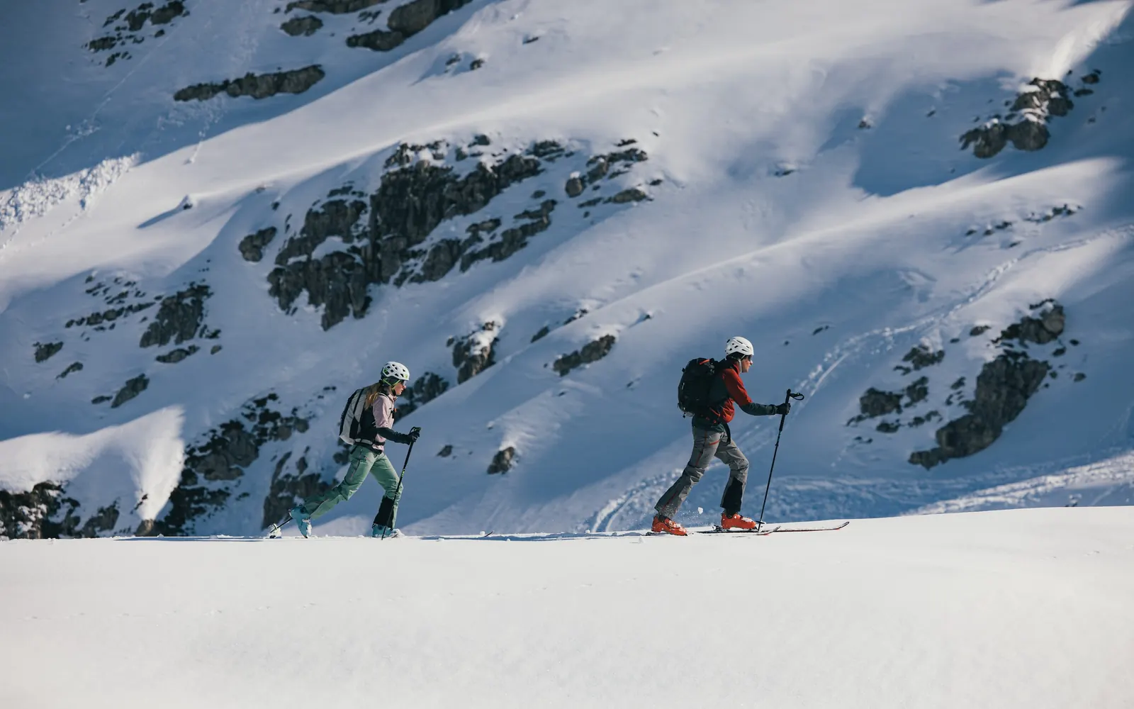 Two ski tourers in Karpos clothing crossing a sunlit snow slope below rocky mountains