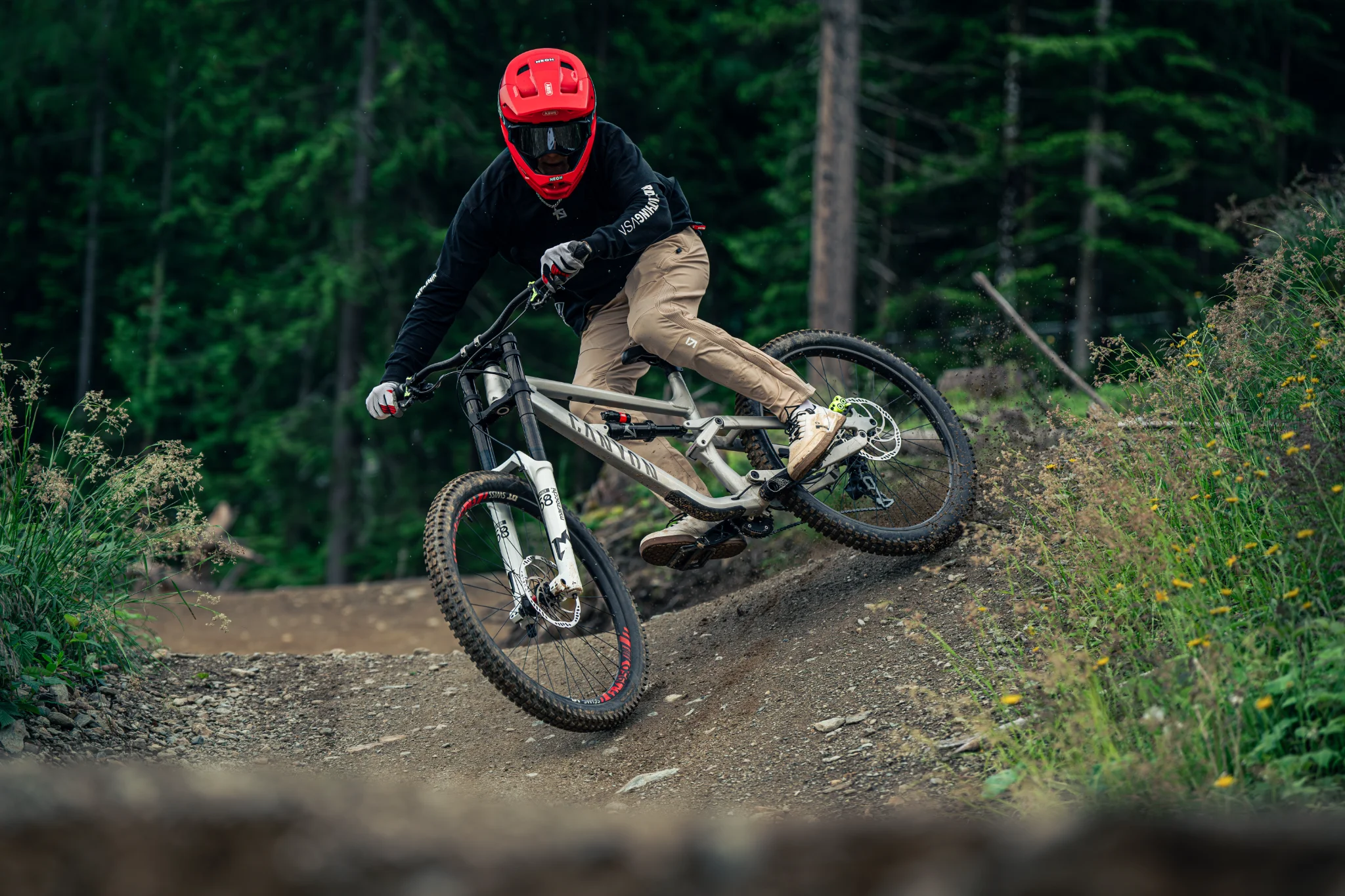 Mountainbiker führt sein Trail-Fully über einen wurzeligen Singletrail im alpinen Nadelwald, Flatpedal-Schuhe klar erkennbar.