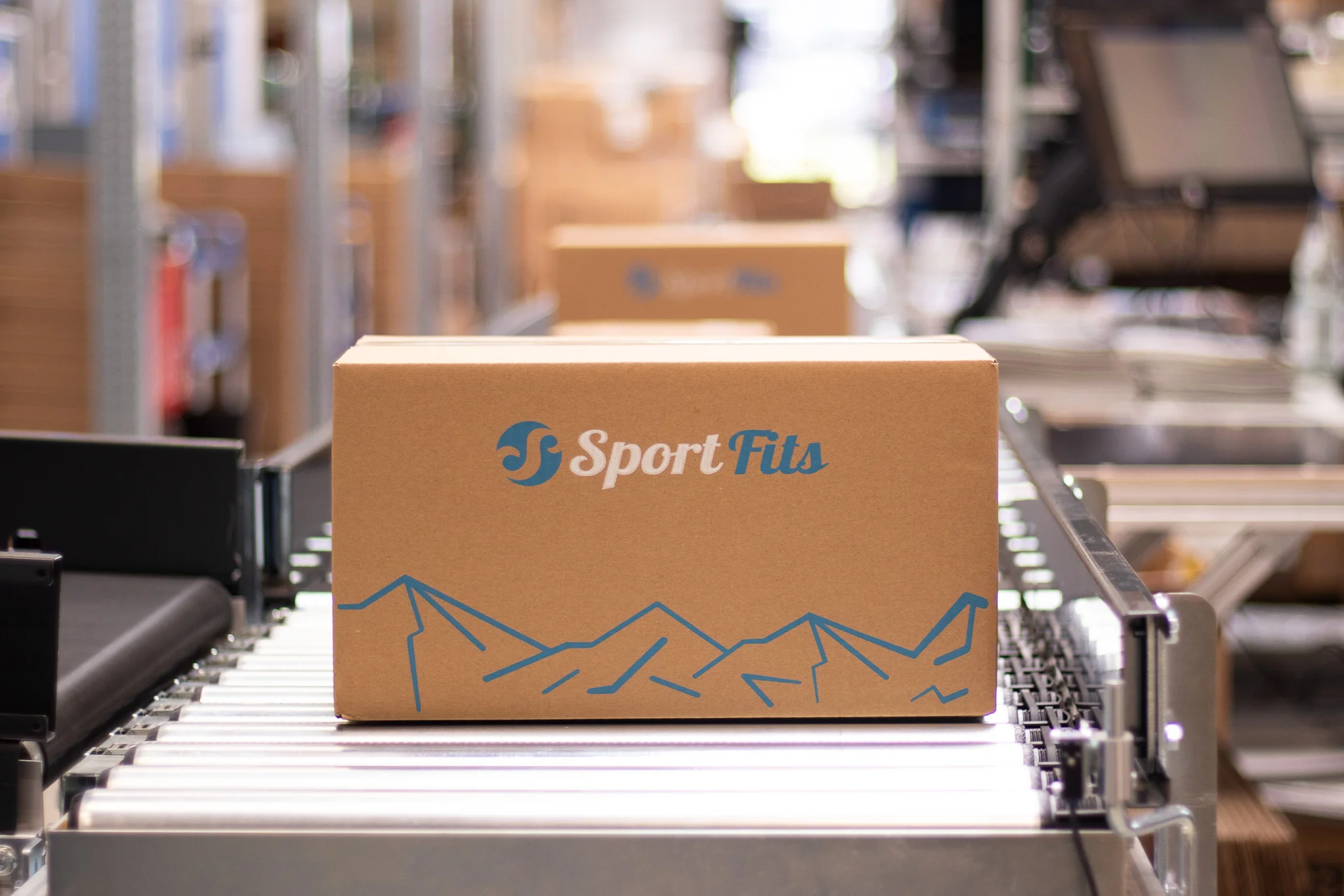 Nahaufnahme eines SportFits-Versandpakets mit Bergmotiv auf einem Foerderband im Logistikzentrum
