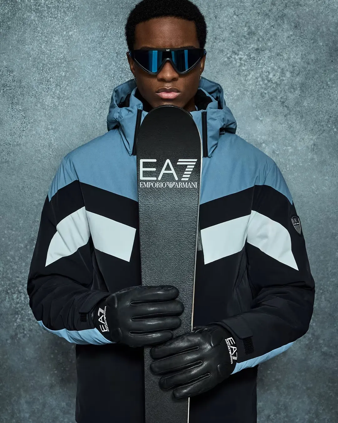 Mann in blau-schwarzer Skijacke mit hellen Streifen und Sportbrille hält einen EA7-Ski vor sich