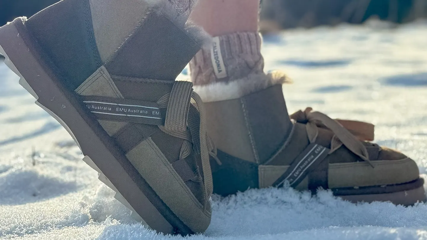 Zwei Schaffell-Stiefel mit Schnürung und EMU-Australia-Band stehen im Schnee, Wintersonne von hinten