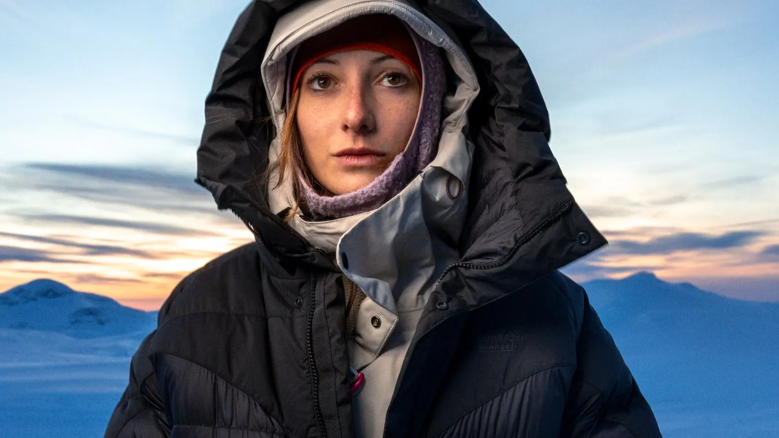 Person mit Kapuze in dunkler, wattierter Jacke vor verschneiten Bergen in der Dämmerung