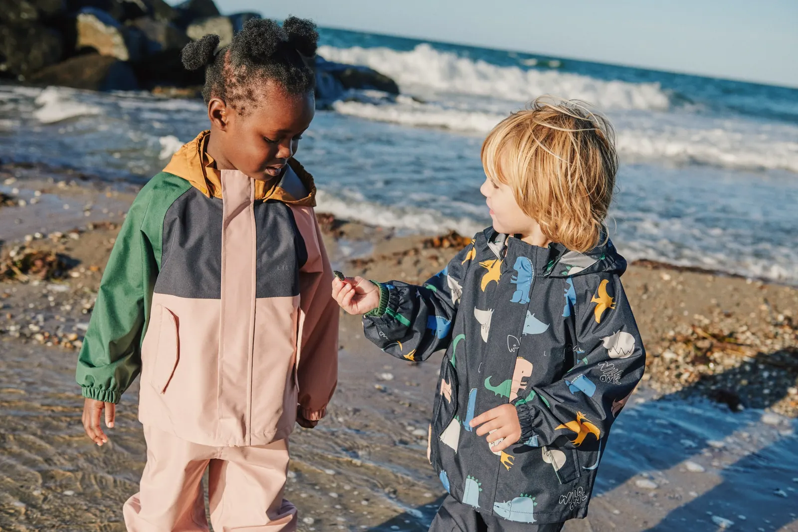 Zwei Kinder in Regenjacken und Regenhosen stehen im flachen Wasser am Strand